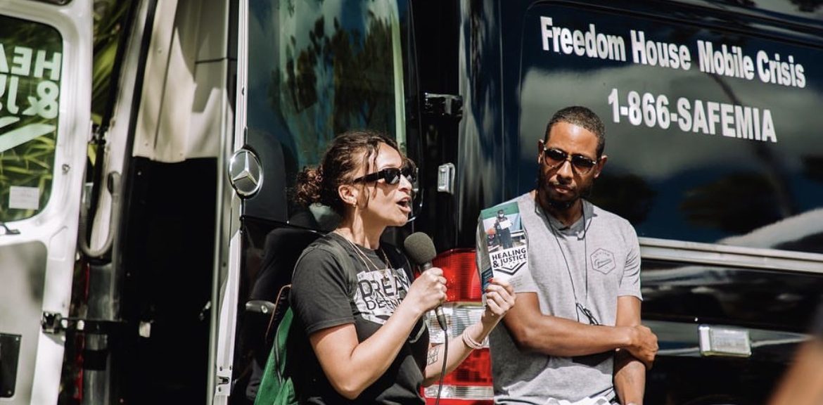 Woman with microphone holding pamphlet next to man outside of a passenger van